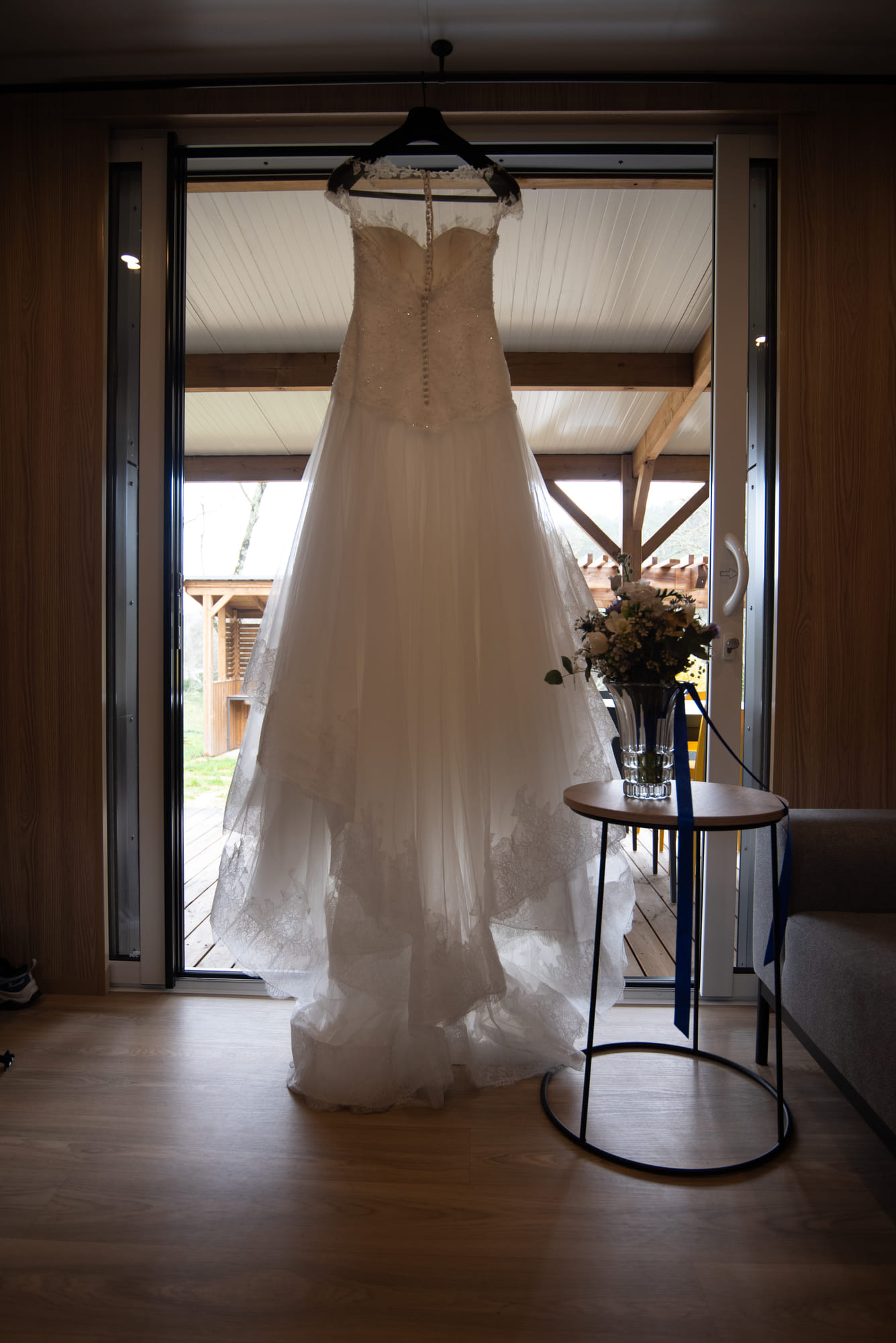 Robe de mariée suspendue devant une baie vitrée, bouquet posé sur une table